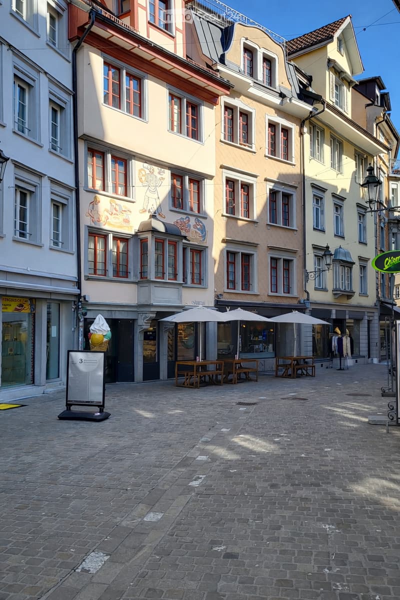 Cobblestone street, multiple buildings, outdoor seating, street lamps, storefronts, mural