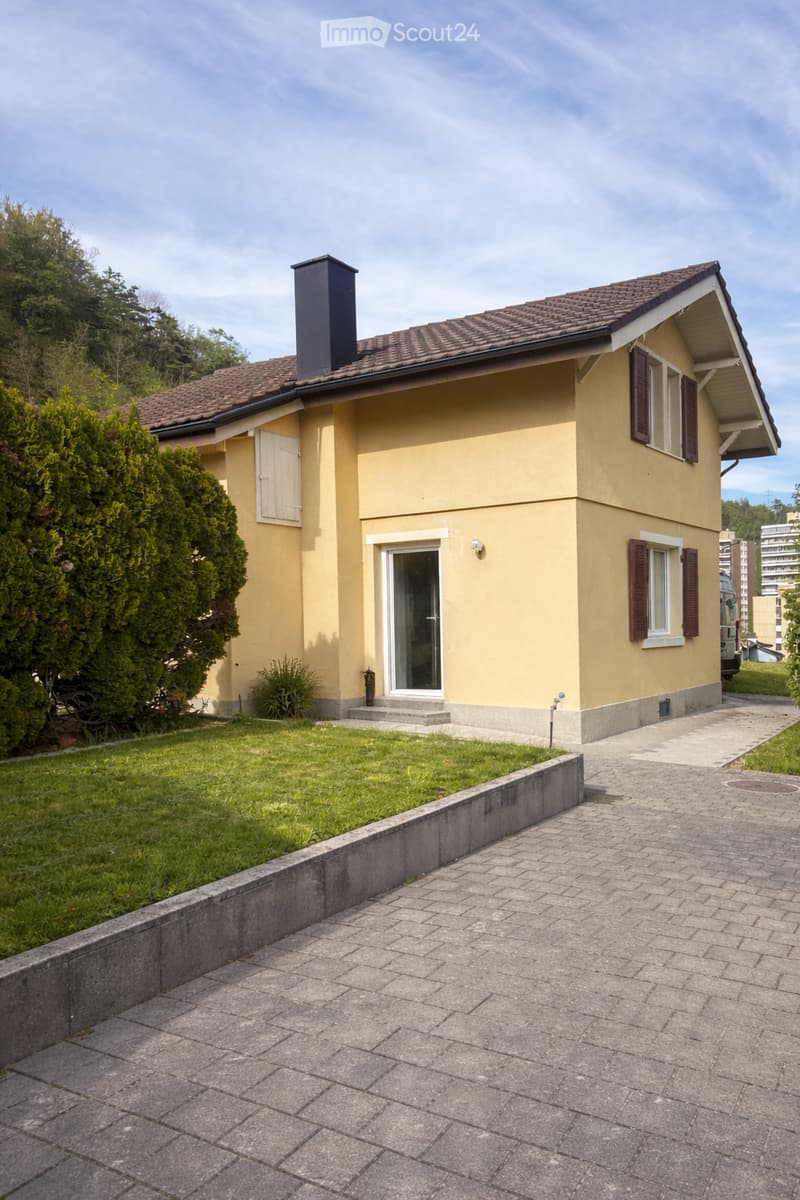 2 story house, yellow exterior, brown roof, glass door, brick driveway, well-maintained lawn, shrubs