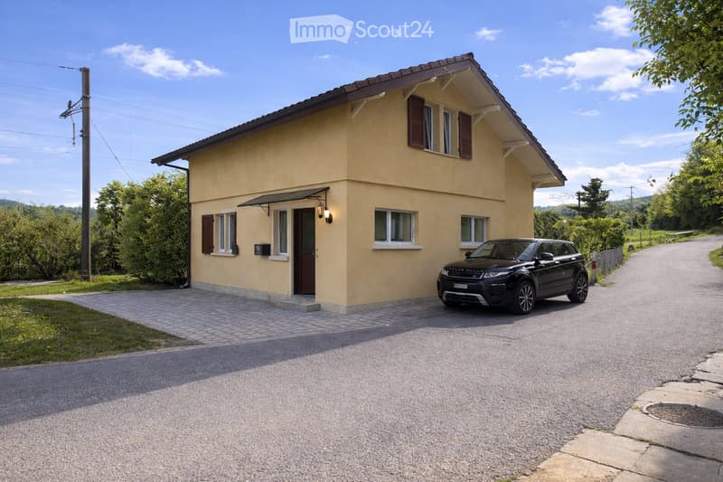 Single-story house, beige exterior, brown roof, driveway, car parked in front, windows with shutters, lamp above the door