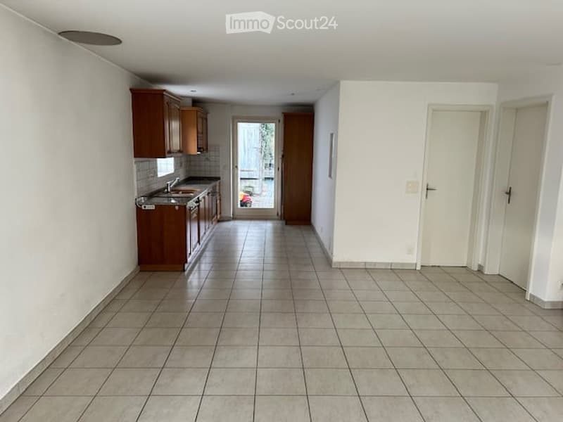 Empty room with white walls, tiled floor, wooden cabinets, sink, and a door leading to another room.