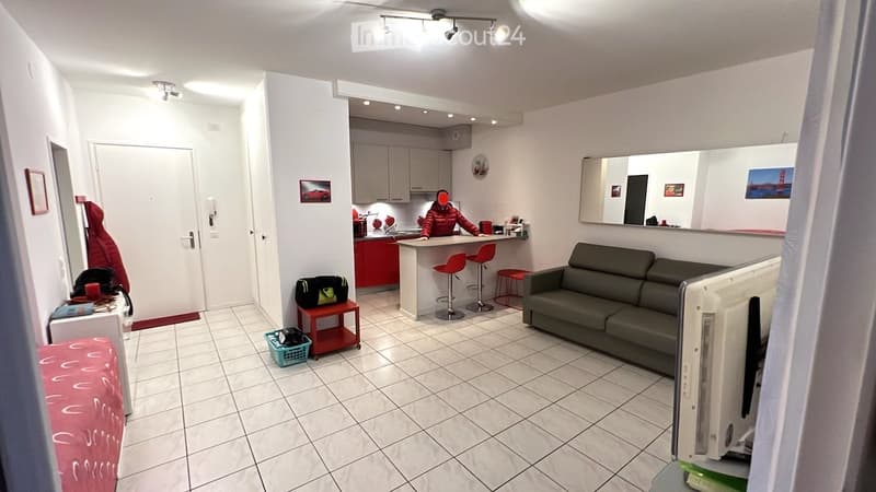 Open plan living room and kitchen, white tiles, gray couch, red stools, kitchen island, mirror, white cabinets, red counter, person in red jacket