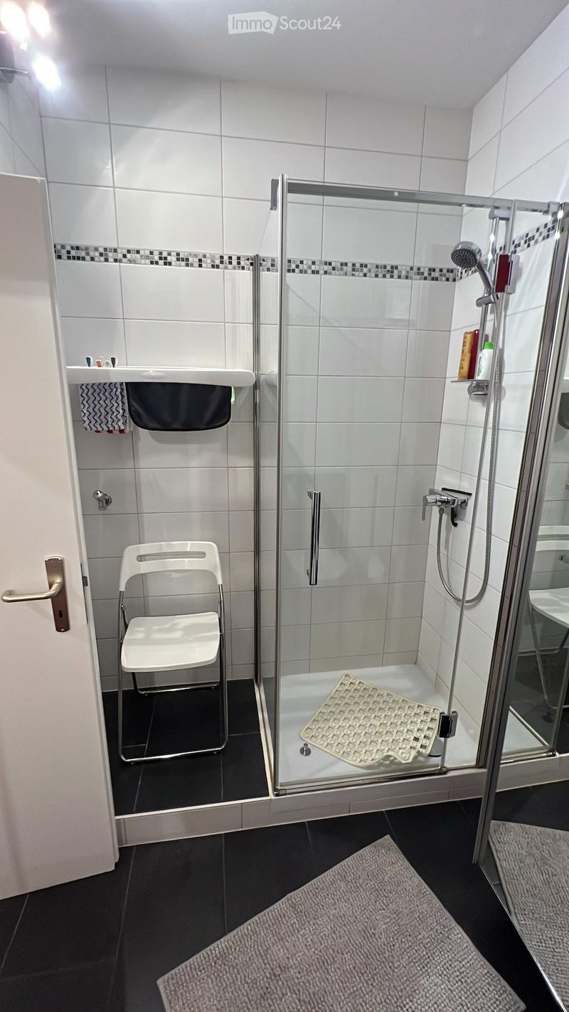 Bathroom with white tiles, glass shower enclosure, white chair, and a rug.