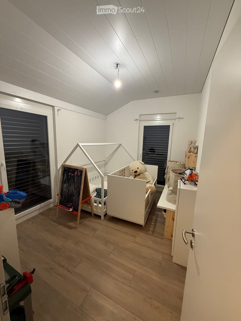 Empty room with wooden floor, white walls, two cribs, blackboard, and toys