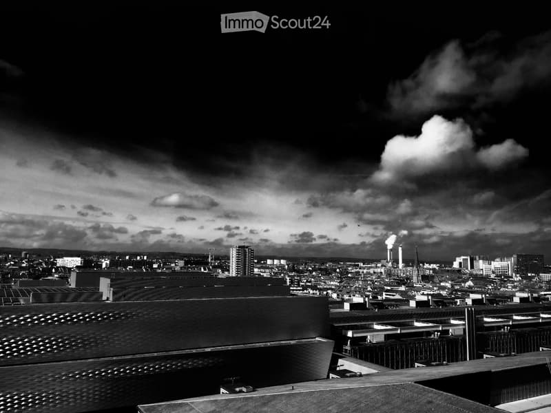 Aerial view of a city with multiple buildings, chimneys emitting smoke, and a cloudy sky.