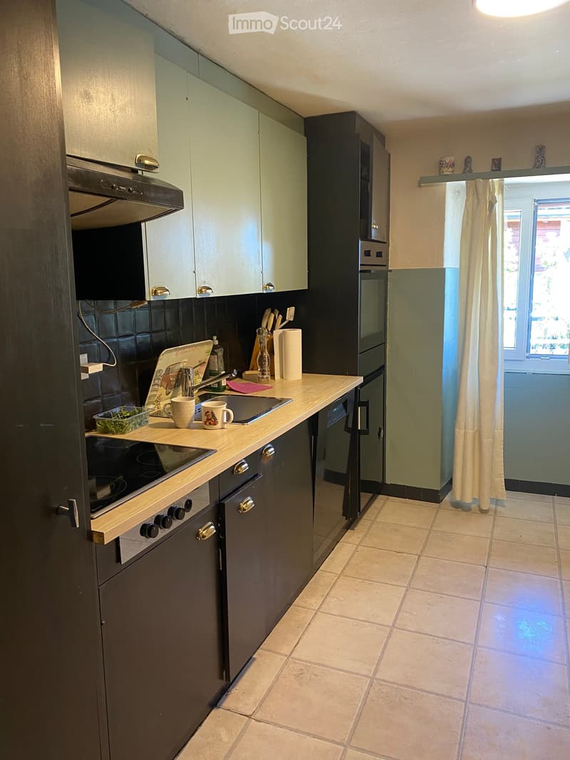 Modern kitchen with light wood countertops, dark grey cabinets, and stainless steel appliances including a stove, oven, refrigerator, and dishwasher. The space features a tiled floor and a window with a curtain.