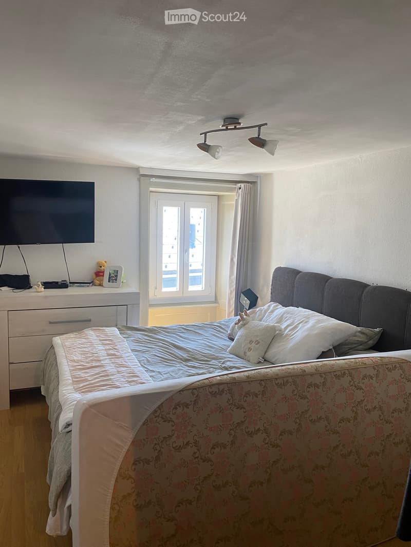 A well-lit bedroom with a large bed, nightstand, lamp, and a window with curtains. The bed has a floral duvet and pillows, and a small nightstand holds a lamp, picture frame, and teddy bear.