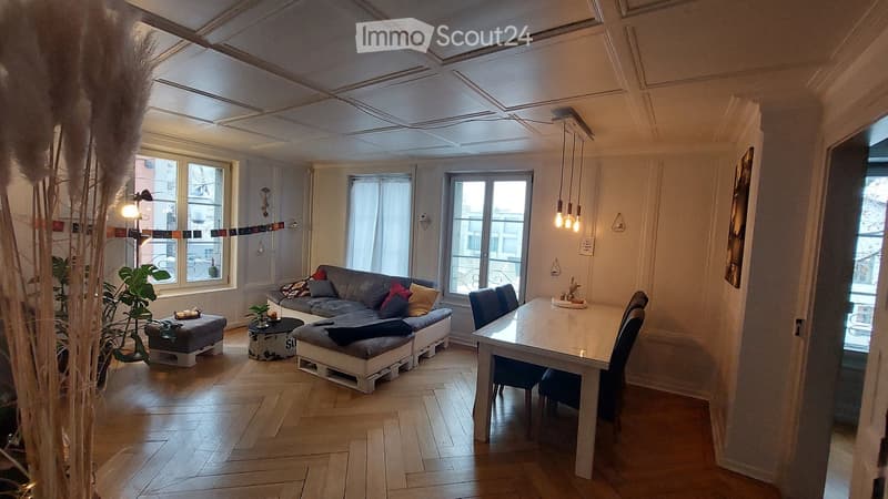 living room with wooden floor, gray sofa, dining table, white walls, large windows, pendant lights, plants