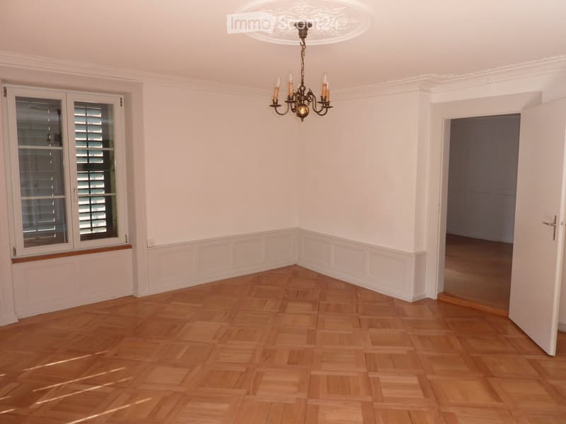 Empty room with wooden parquet floor, white walls, chandelier, window with shutters, and an open door leading to another room.