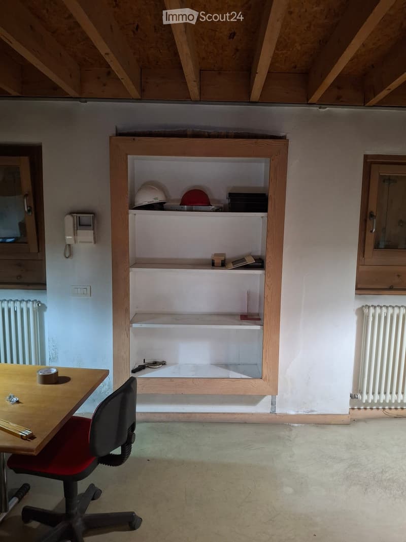 A small, unfurnished room with a wooden shelf containing helmets and tools, a table with a chair, and exposed wooden beams on the ceiling.
