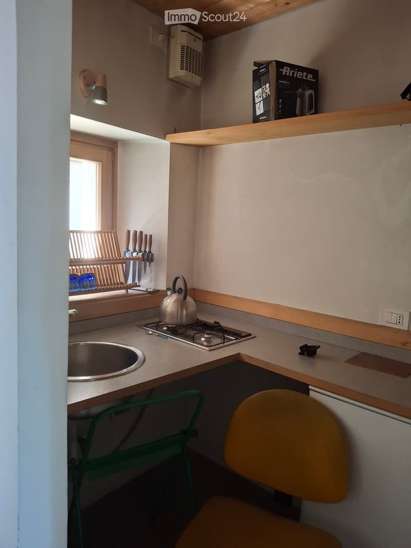 Compact kitchen with a sink, stove, teapot, knife rack, and a small table with two chairs. Wooden shelves and a wall-mounted lamp are visible.