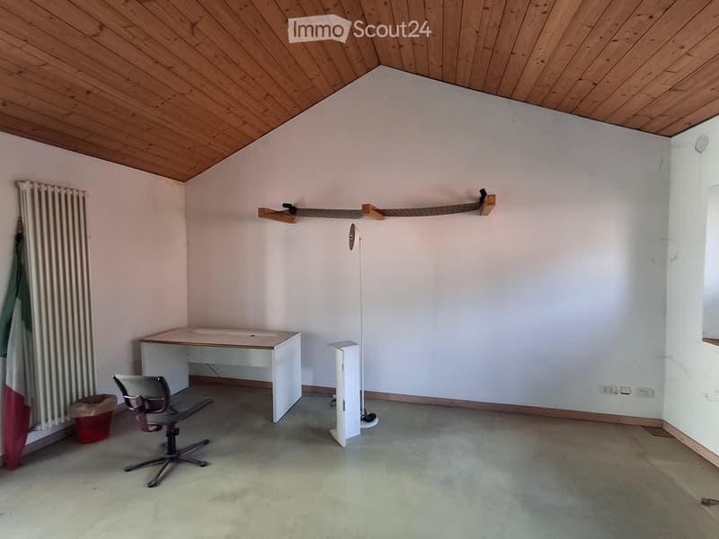 A small, unfurnished room with a wooden ceiling, a desk, a chair, a radiator, and a shelf. The floor is concrete, and the walls are white.