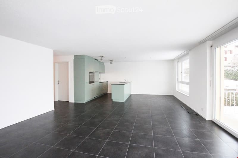 Empty room with white walls, black tiled floor, kitchen with countertop, cabinets, and appliances, sliding glass door leading to balcony