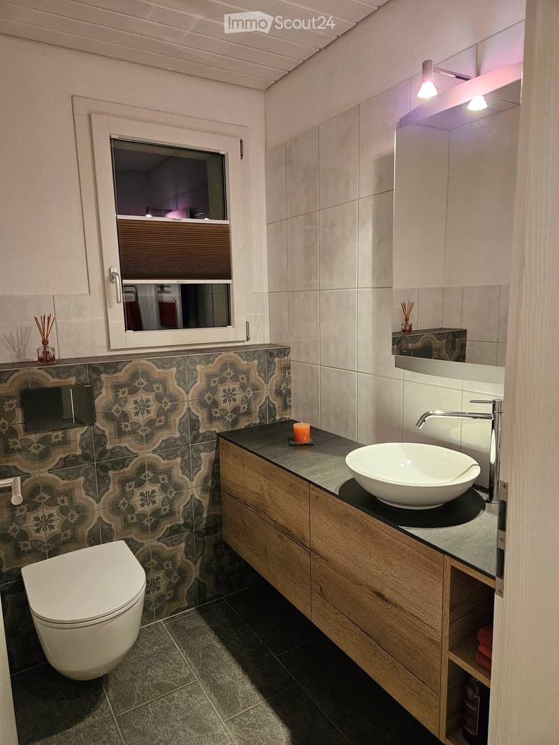 White toilet, decorative tiles, white countertop, modern sink, wooden cabinetry, under-mount sink, mirror with lighting, modern fixtures