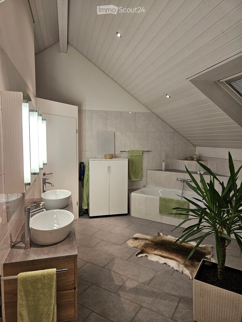 modern bathroom with wooden cabinets, two sinks, bathtub, wall mounted fixtures, white ceiling, tiles, rug, potted plant, towel racks