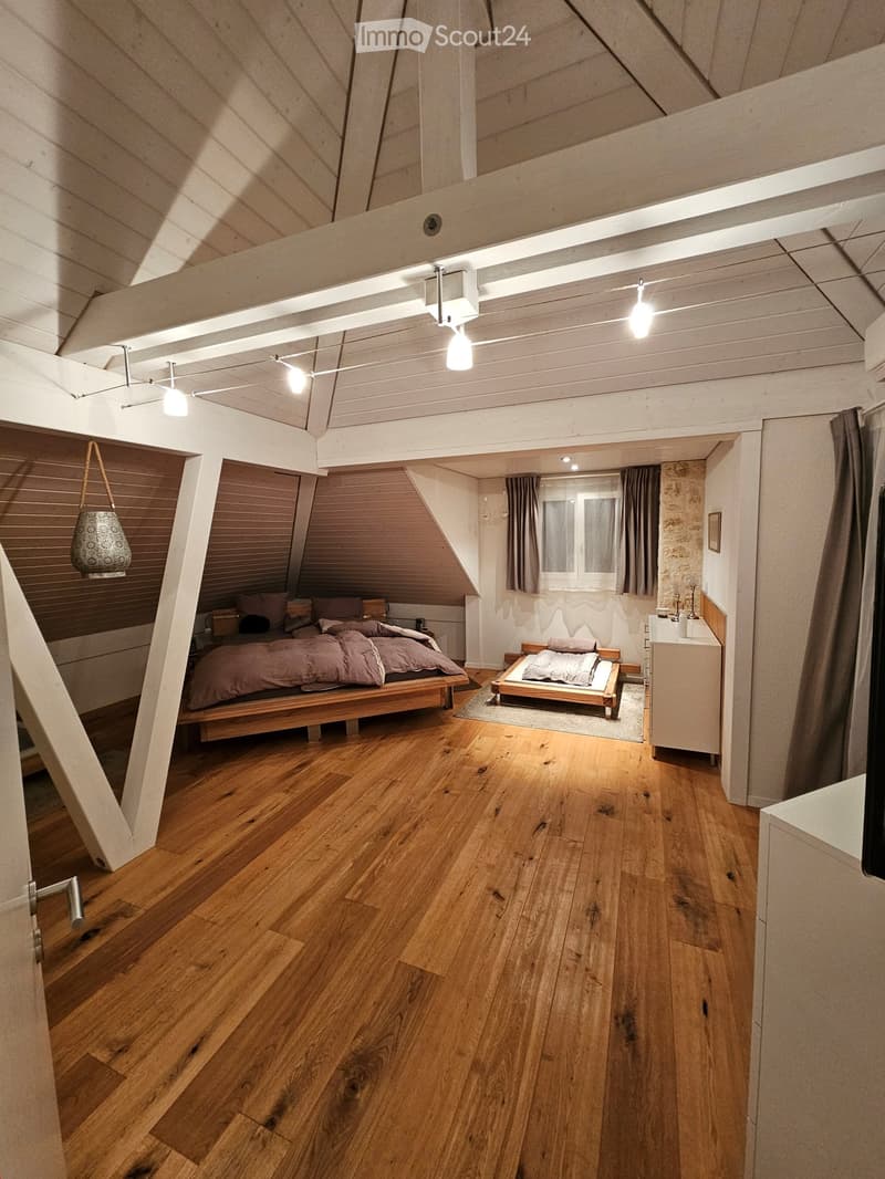Two beds, one large and one small, both with linen, on wooden frames, wooden floor, ceiling with cross beam and lights, small window with curtains, dresser.