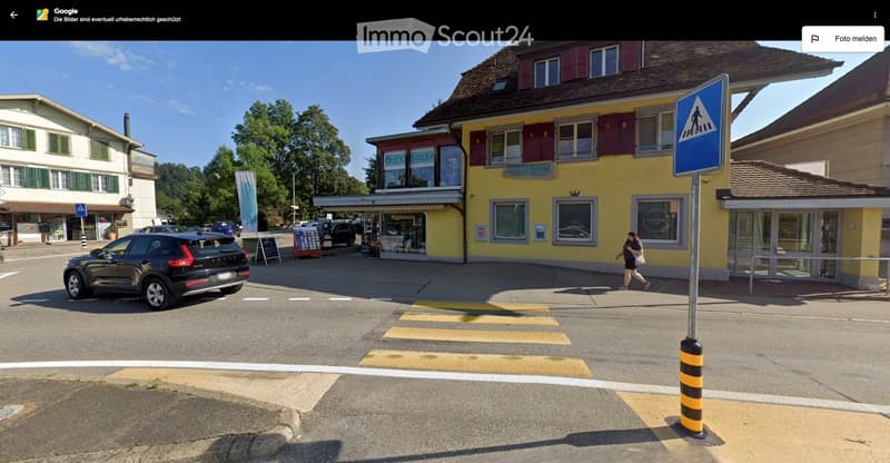 A street view of a building with a yellow facade and red shutters, a pedestrian crossing sign, and a car parked on the side of the road.