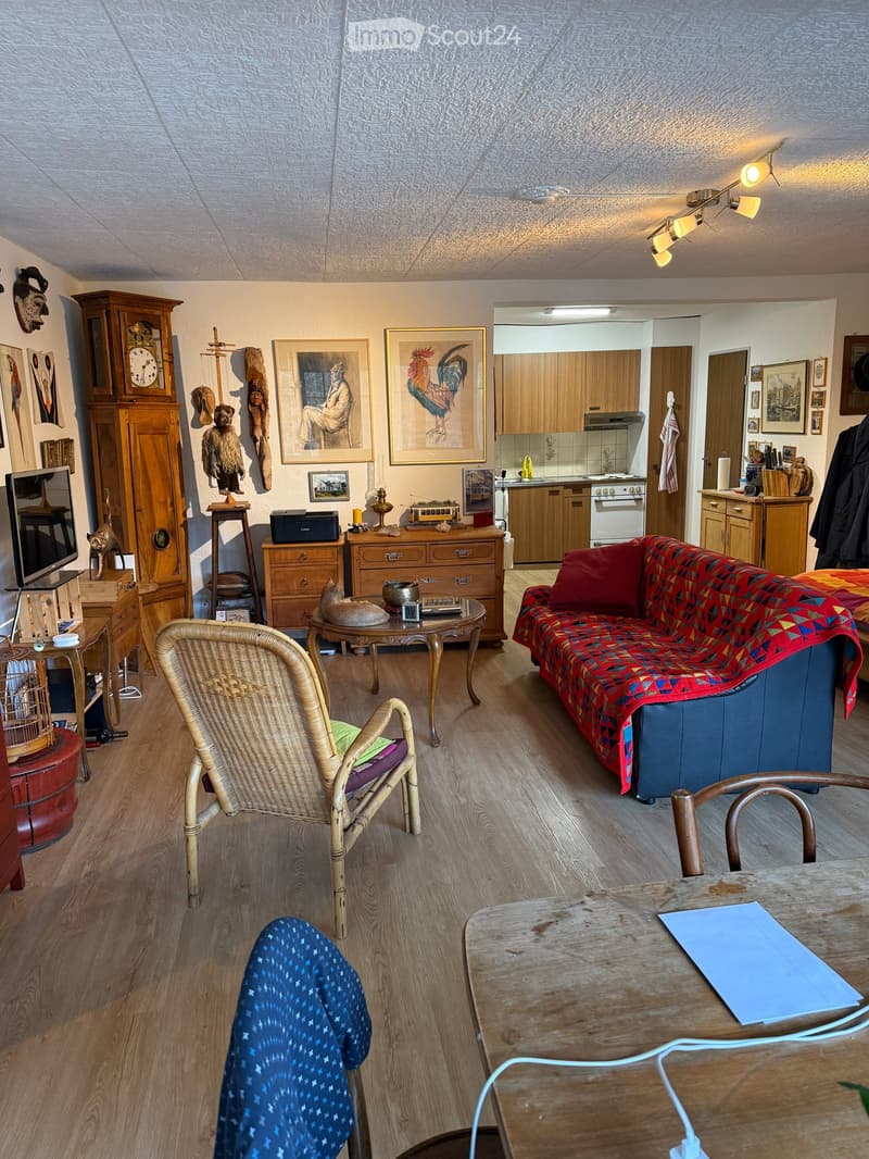 Living room with wooden floor, wooden furniture, a couch, a table, a chair, a television, a cabinet, a clock, and a kitchen area.