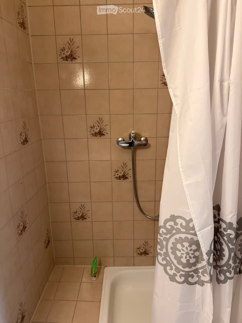 small shower area with tiled walls, shower head, shower curtain, small bathtub, green bottle