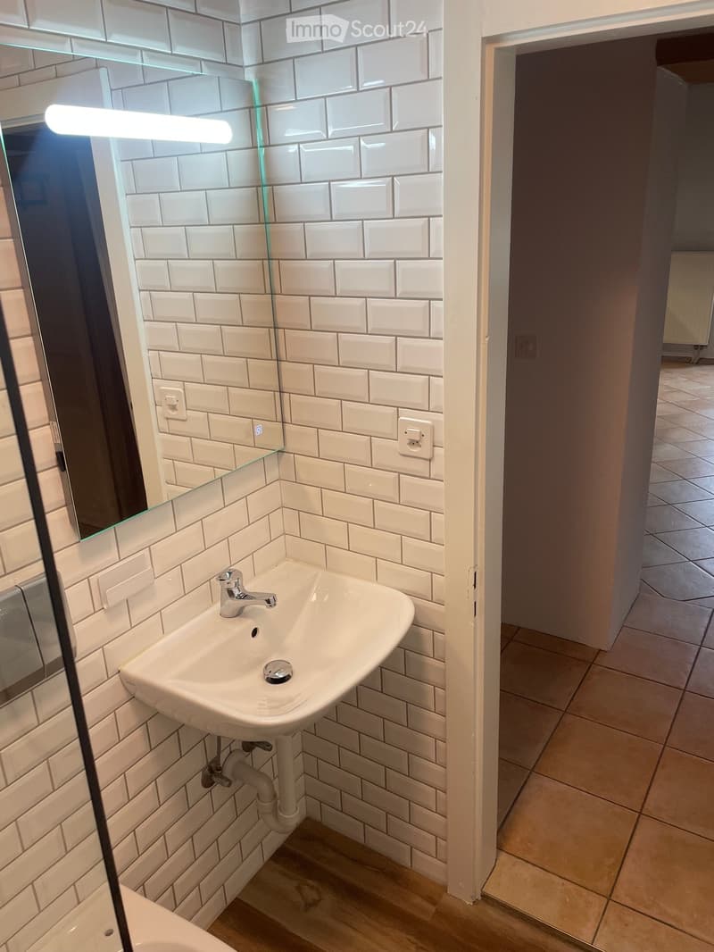 A small bathroom with white tiled walls and tiled floor, a sink with faucet, a mirror, and a toilet.