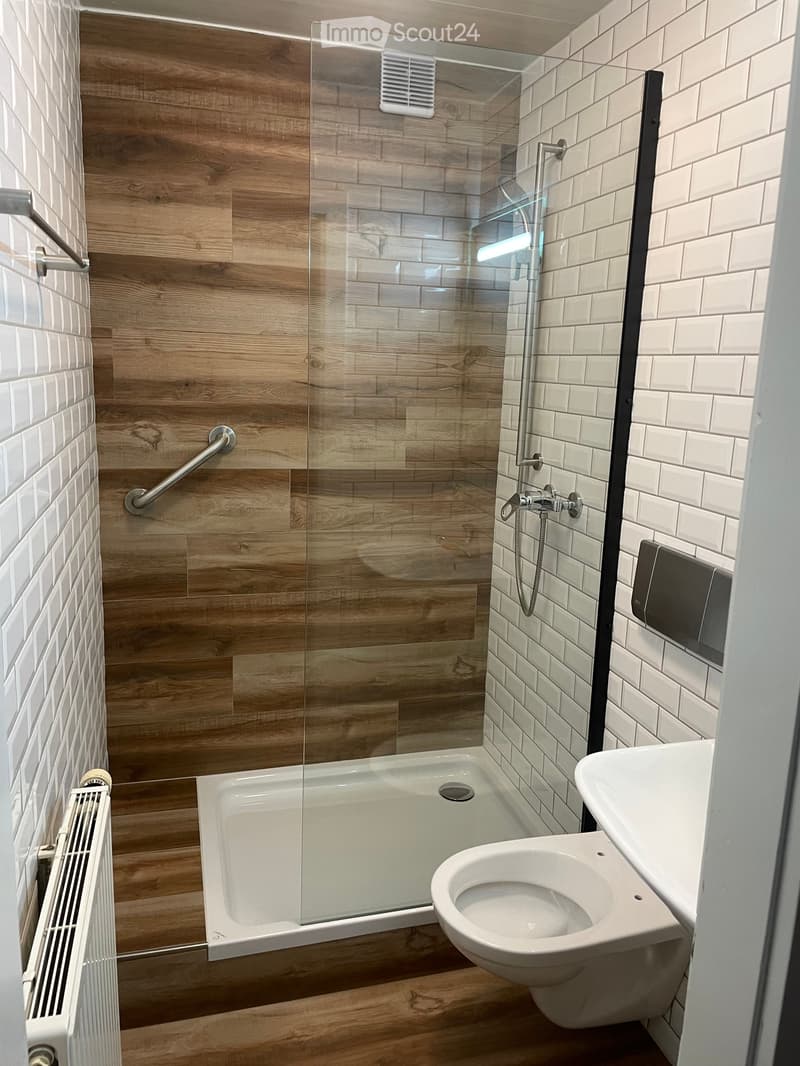 A modern and minimalistic bathroom with wooden flooring, white tiled walls, a glass shower enclosure with chrome fixtures, a white toilet bowl, a white sink, and a towel rack.