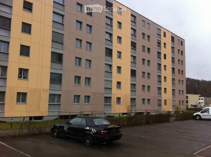 A multi-story residential building with yellow and brown exterior walls, multiple windows, and a parking area in front.