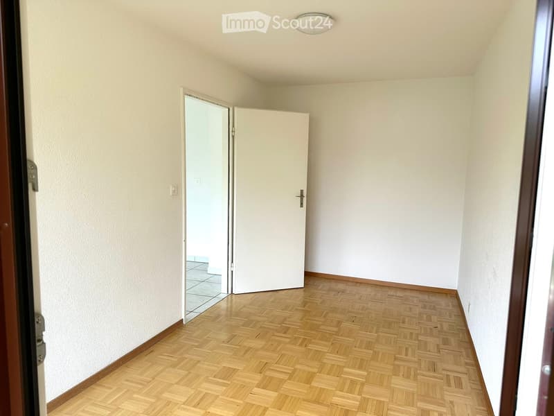 Empty room with white walls, wooden parquet flooring, white door, and an open door leading to another room with tiled flooring.