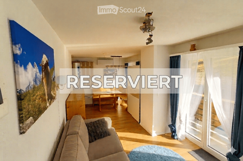 Bright living room with wooden flooring, a sofa, a coffee table, a dining area with a table and chairs, a large picture frame on the wall, and a sliding glass door leading to a balcony.