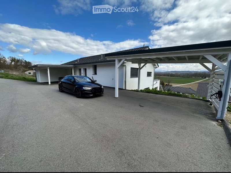 white house, black car parked under a carport, driveway, green field in the background