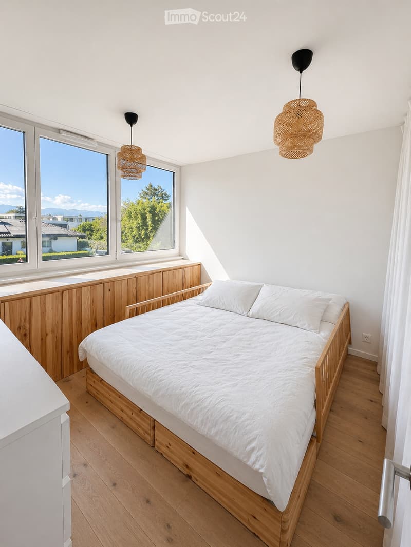 Bedroom with wooden bed, white linens, wooden window seat, two lamps, wooden floor, white walls, large windows with view