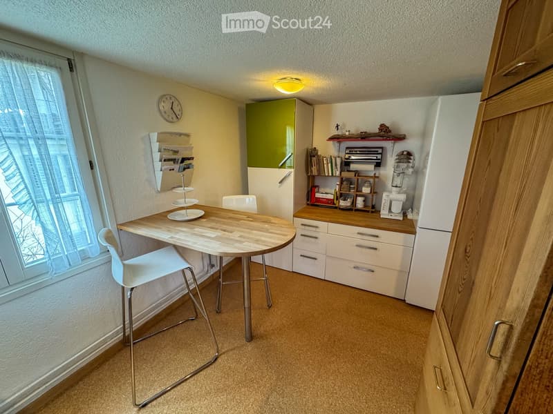 small room with a table, chairs, a window, a clock, a shelf, a refrigerator, and a cabinet