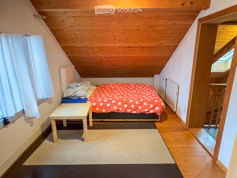 A small bedroom with a bed, a small table, a carpet, and a window with white curtains.