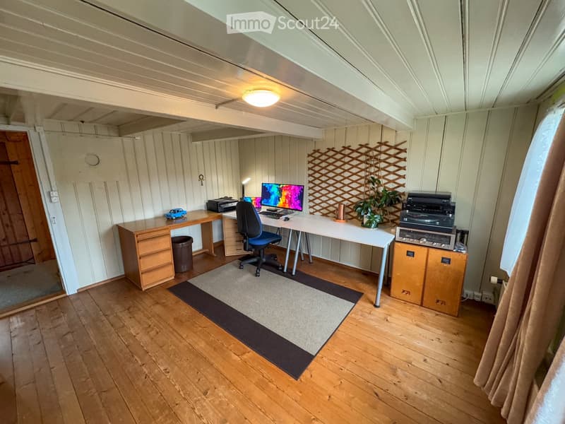 A room with wooden floors, white walls, and a ceiling with a ceiling lamp. There are two desks with monitors, a printer, and a chair. There is a rug on the floor, a potted plant, and a speaker. There is a wooden cabinet with a turntable and a power outlet