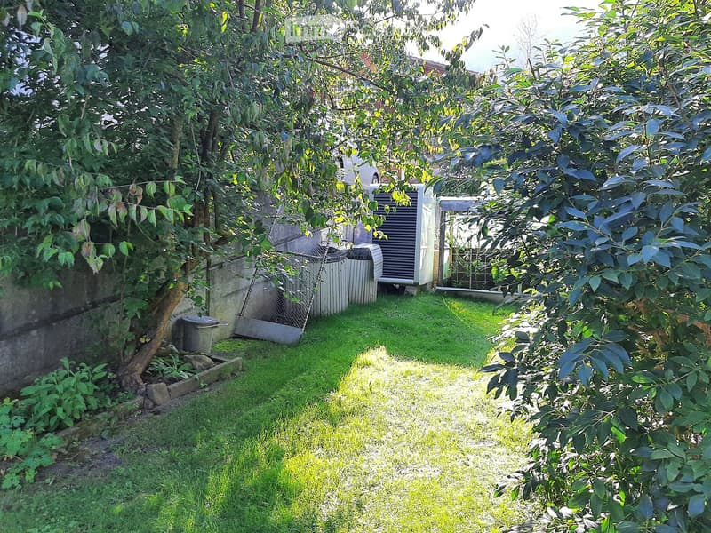 A small backyard with a grassy lawn, a tree, and a wall. There are some plants and a trash bin on the left side. A white air conditioner is placed on the wall. A metal fence is on the right side, and a building is behind it.