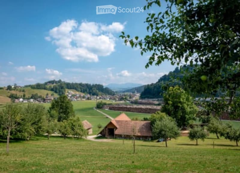 Scenic rural landscape with rolling hills, green fields, a traditional barn or farmhouse, and a village in the distance surrounded by trees and mountains.