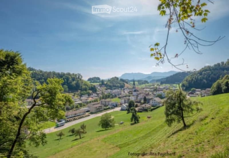 Scenic view of a small town nestled in a valley surrounded by lush green hills and forests. The town has a church steeple visible in the center. The landscape is characterized by rolling hills, trees, and a clear blue sky with some clouds.