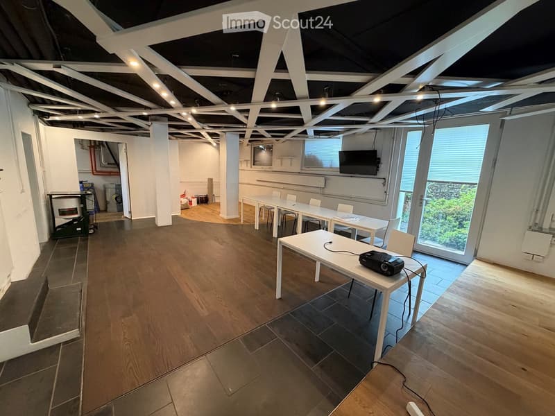 Open-plan room with wooden flooring, white walls, exposed ceiling beams, and large windows. Contains tables, chairs, and a projector on one table.