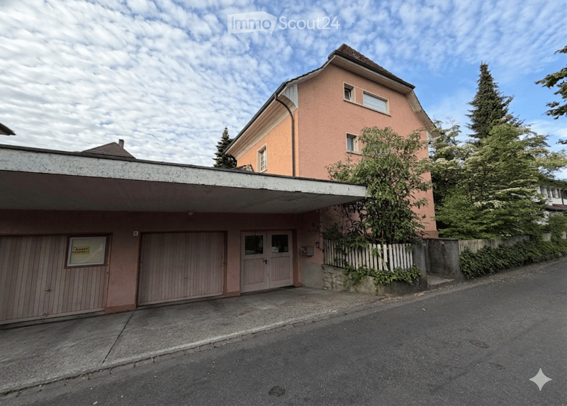 Two-story building with a garage, pink facade, and multiple windows. The entrance features a double door and a covered area.