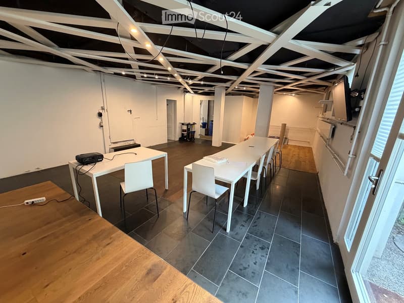 Bright, open room with exposed ceiling beams, two white tables with chairs, and a screen on the wall. The floor is a mix of dark tiles and light wood.
