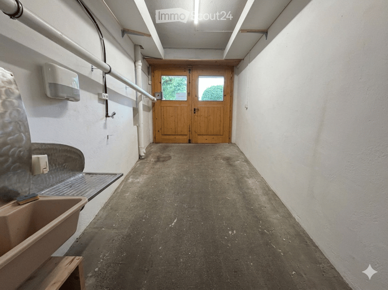 A narrow, concrete-floored hallway with white walls, a wooden double door at the end, and a small sink area on the left side.