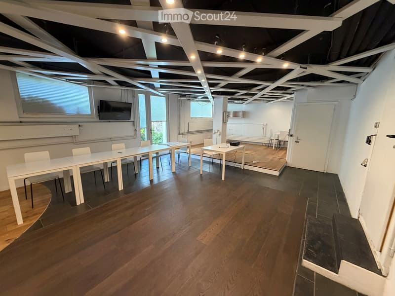 Bright, open room with wooden flooring, white tables, chairs, and a mounted screen. Exposed ceiling beams and large windows provide natural light.