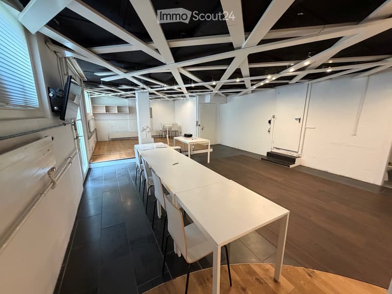Open-plan space with exposed wooden beams and modern lighting, featuring a long white table with chairs, a smaller table with chairs, and a TV screen mounted on the wall.