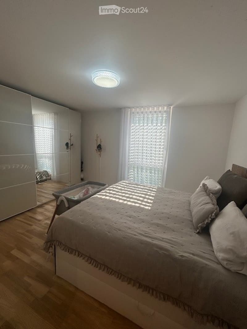 Spacious bedroom with a bed, crib, and a wardrobe. The room has a white ceiling, wooden floor, and white walls. There is a lamp on the ceiling and a crib in the corner.