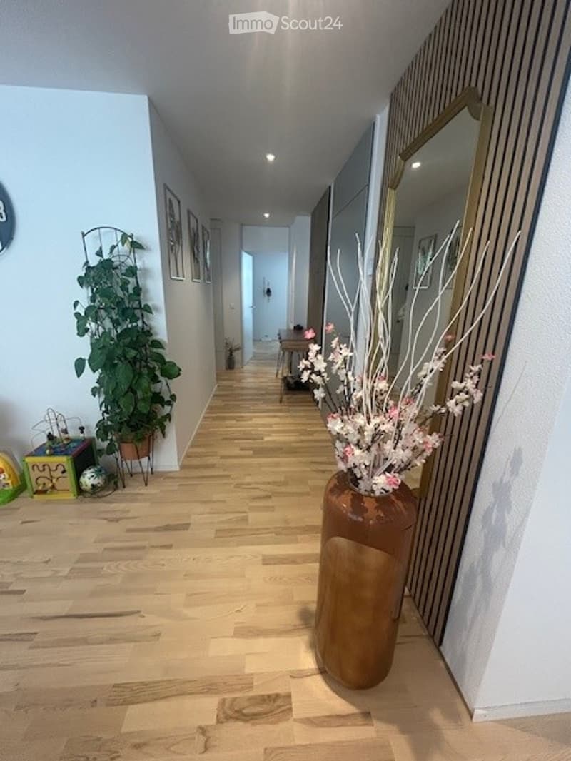 A hallway with wooden flooring, a large vase with flowers, a potted plant, a mirror, and picture frames on the wall. There is a table and chairs in the distance, along with some toys on the floor.