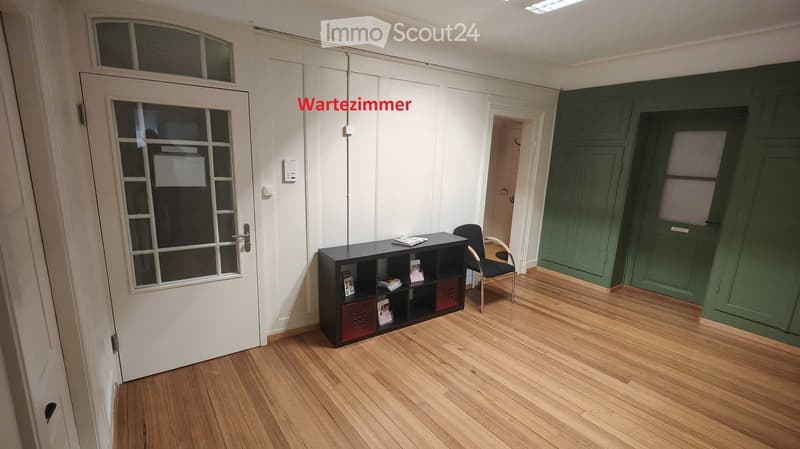 An empty room with wooden floors, white walls, and green doors. A black shelf with books and a chair is placed against the wall. The room has a glass door and a window.