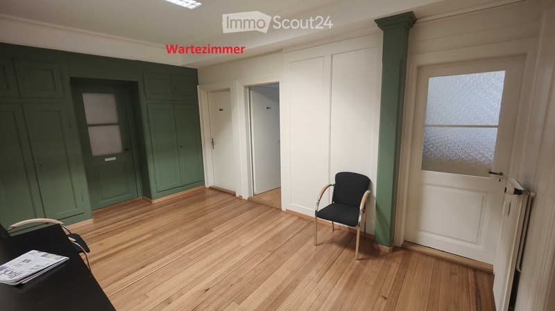 An empty room with wooden floors, green walls, and white doors. There are two black chairs and a desk with a newspaper on it.
