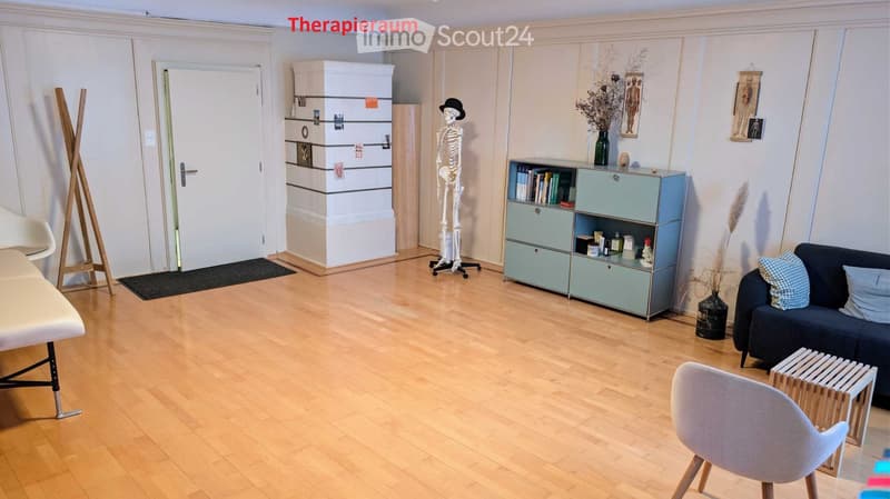 Spacious room with wooden floor, white walls, and various furniture. A skeleton stands in the center, a cabinet with books and a vase is on the right, and a couch with pillows is on the right side. A chair and a small table are on the right, and a stand w