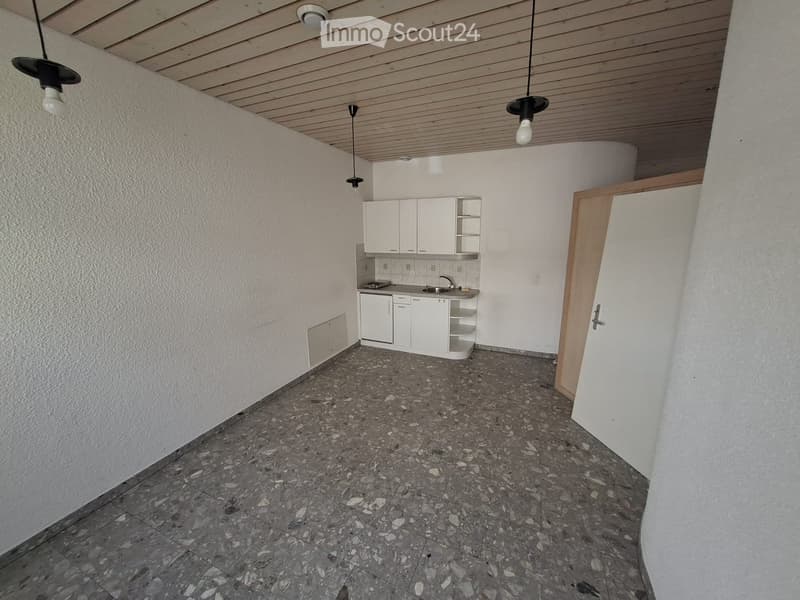 Empty room with white walls, wooden ceiling, and gray tiled floor. There are white cabinets, a sink, and a faucet in the corner. There are three hanging lamps.