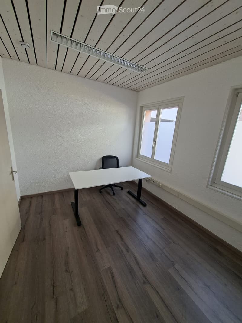 An empty room with a white desk, a black office chair, and two windows. The room has a wooden floor and white walls.