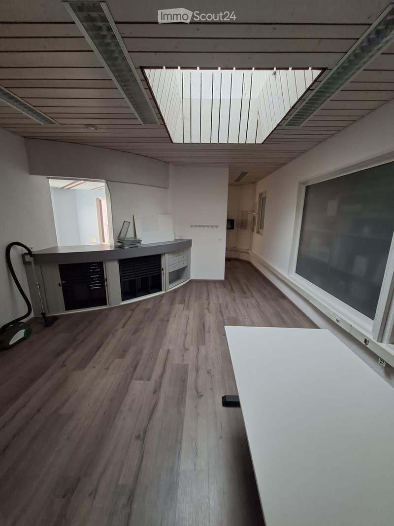 Empty room with wooden floor, white walls, and a large window. A desk and a vacuum cleaner are present.