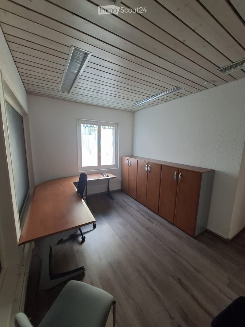 An empty room with wooden floors, white walls, and a window. There are two desks, one with a chair, and a set of wooden cabinets against the wall.